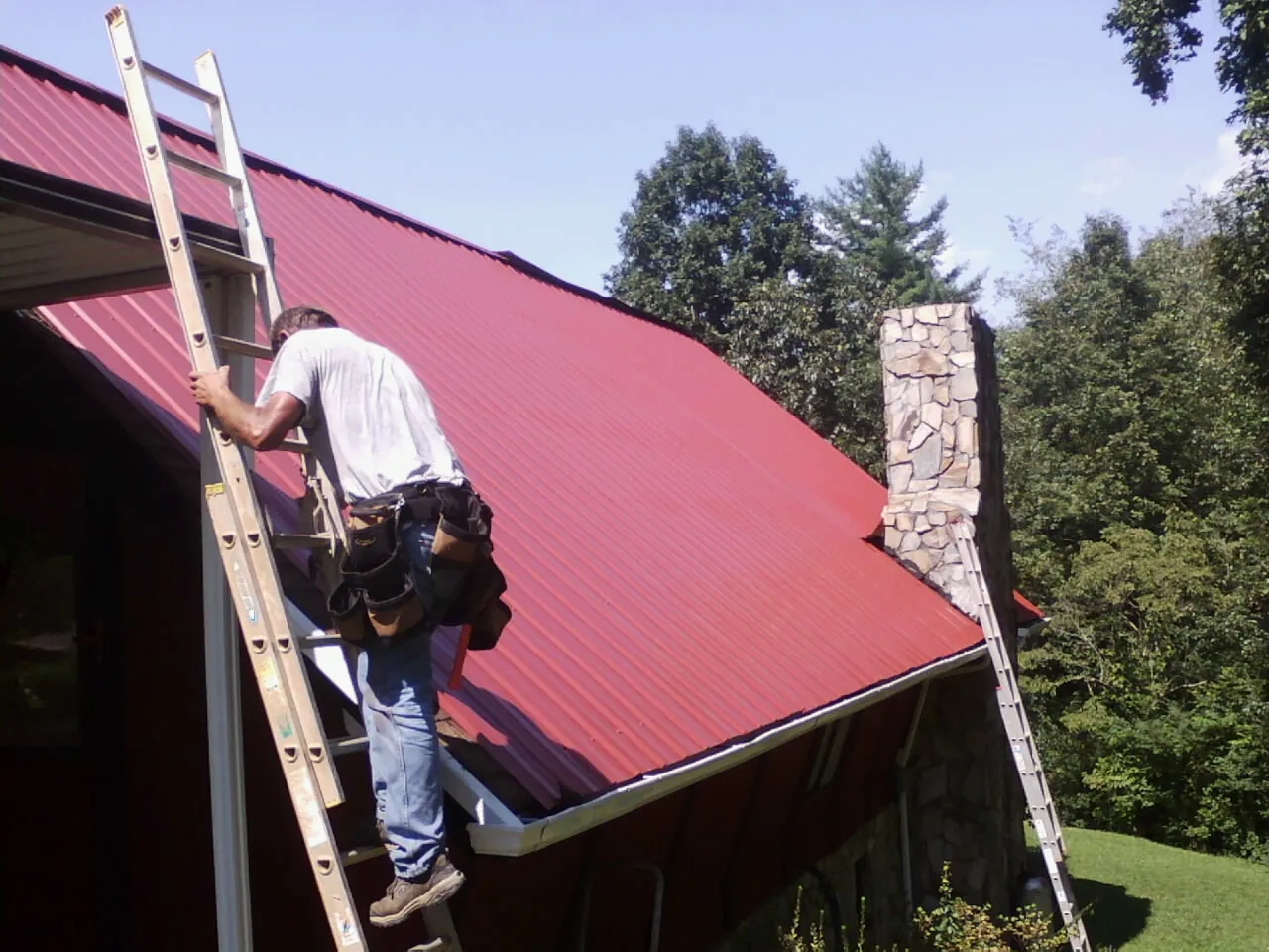 Roofer on a ladder beside a red metal roof and stone chimney — Soffit & Fascia Repair by Roofing Brothers in Pulaski